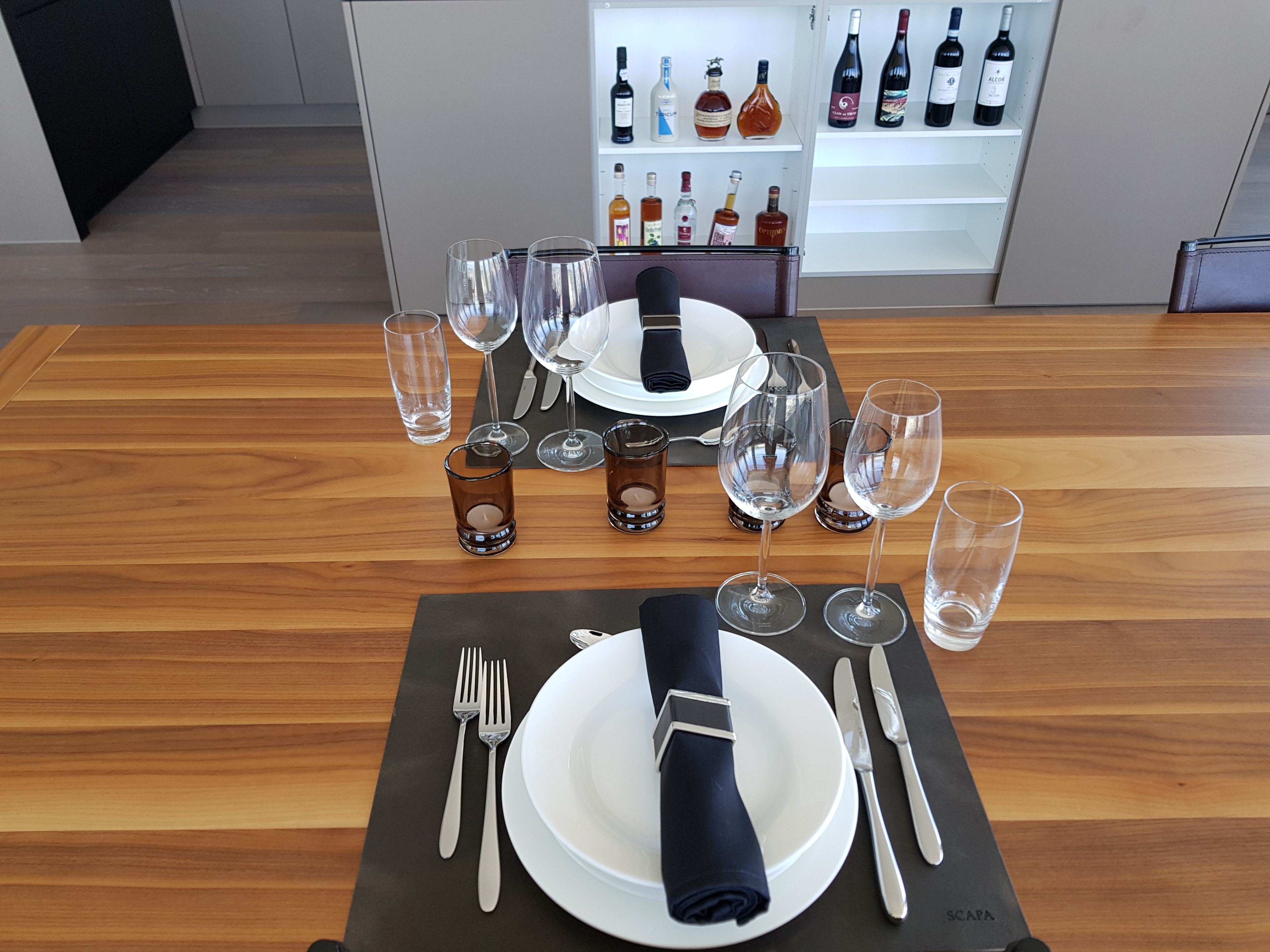 A dining table set with wine glasses, water glasses, and cutlery, with a wine refrigerator in the background.
