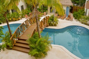 Un área de piscina con un puente de madera, sillas de descanso y plantas tropicales.