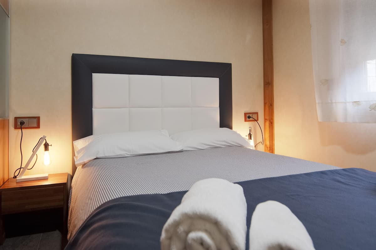 A neatly made bed with a headboard, two bedside tables with lamps, and a window with sheer curtains.