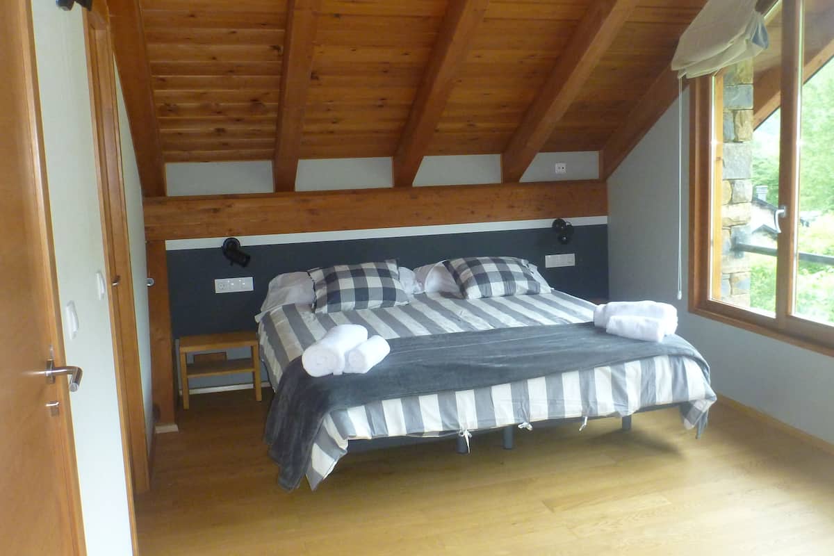 A bedroom with a wooden ceiling, a bed with striped bedding, and a window with a view of greenery outside.