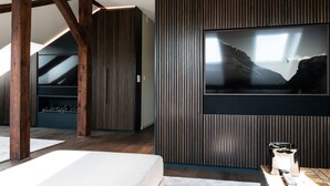 A modern room with a large TV mounted on a wall, wooden beams, and a stone fireplace.