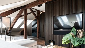 A modern living room with a large flat-screen TV mounted on a dark wall, wooden beams, and a large plant on a wooden coffee table.