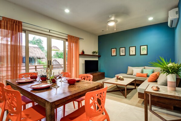 A dining area with a wooden table, orange chairs, and a TV. There is a sofa with cushions and a plant on a side table.