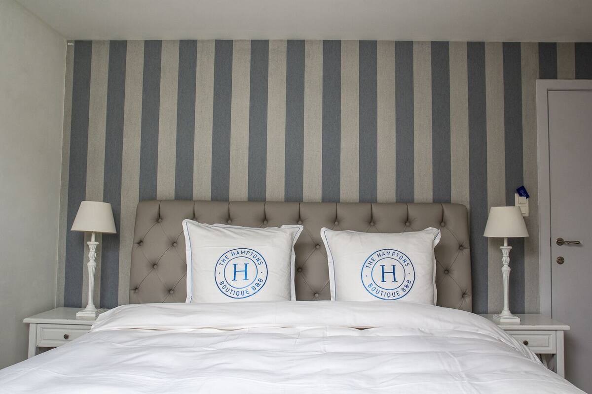 Une chambre avec un mur à rayures, une tête de lit capitonnée, deux tables de chevet avec des lampes et deux coussins ornés d’un logo.