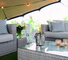 A wicker sofa with patterned cushions, a glass table with a champagne bucket and glasses, and decorative lights.