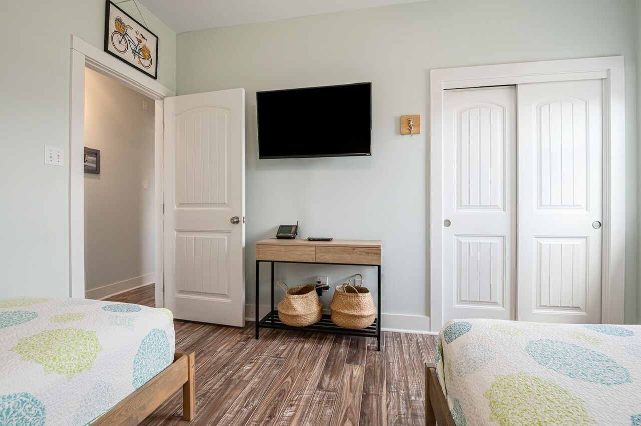 A bedroom with two beds, a TV mounted on the wall, a wooden desk with baskets, and white doors.