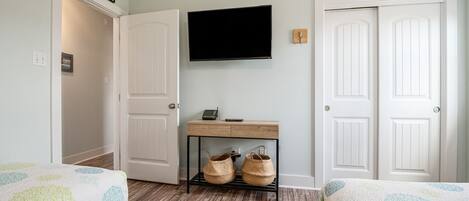 A bedroom with two beds, a TV mounted on the wall, a wooden desk with baskets, and white doors.