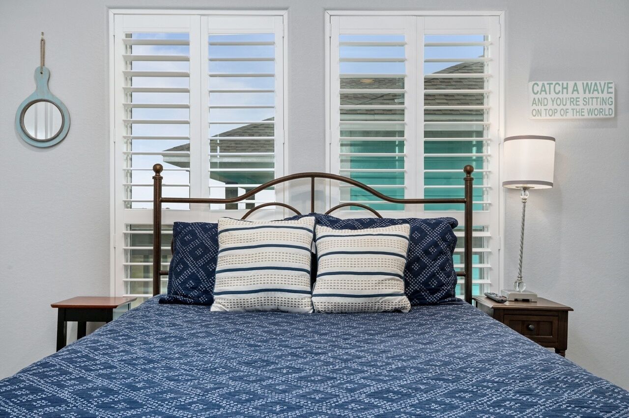Un dormitorio con cama, dos mesitas de noche, una lámpara de pared, un espejo colgado y una ventana con persianas.