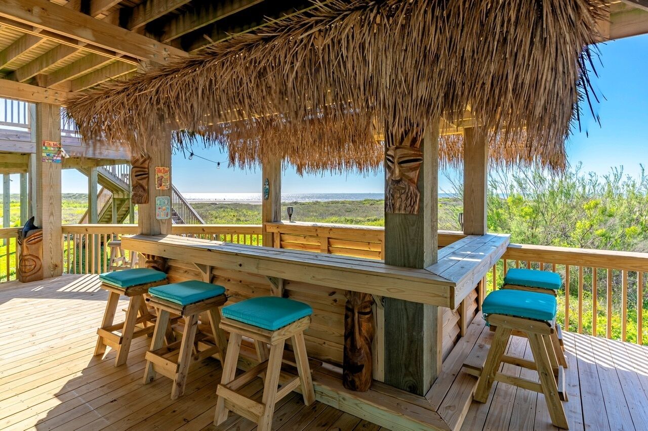 A wooden deck with a thatched roof, wooden bar, and stools.