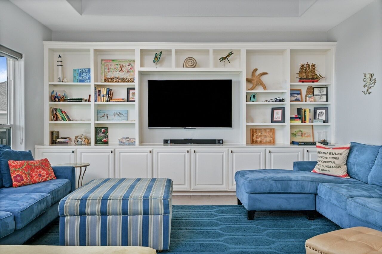 A living room with a large flat-screen TV, blue sofas, a striped ottoman, and a bookshelf with various decorative items.