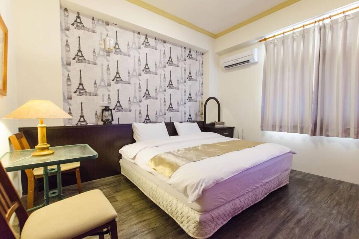 A hotel room with a bed, a desk with a lamp, a chair, a mirror, and a window with curtains.