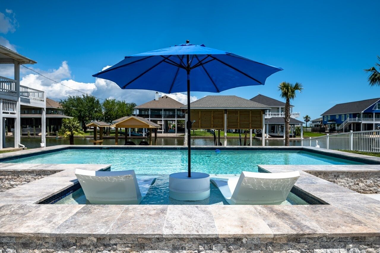 Una piscina con un paraguas azul, dos sillas de descanso blancas y una terraza de piedra.