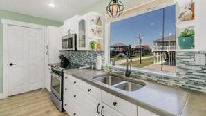 Une cuisine avec des meubles blancs, un évier à deux bacs, un four à micro-ondes et une fenêtre donnant sur une maison et un balcon.