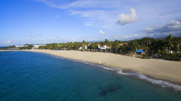 A beach with clear blue water, palm trees, and buildings along the shore.
