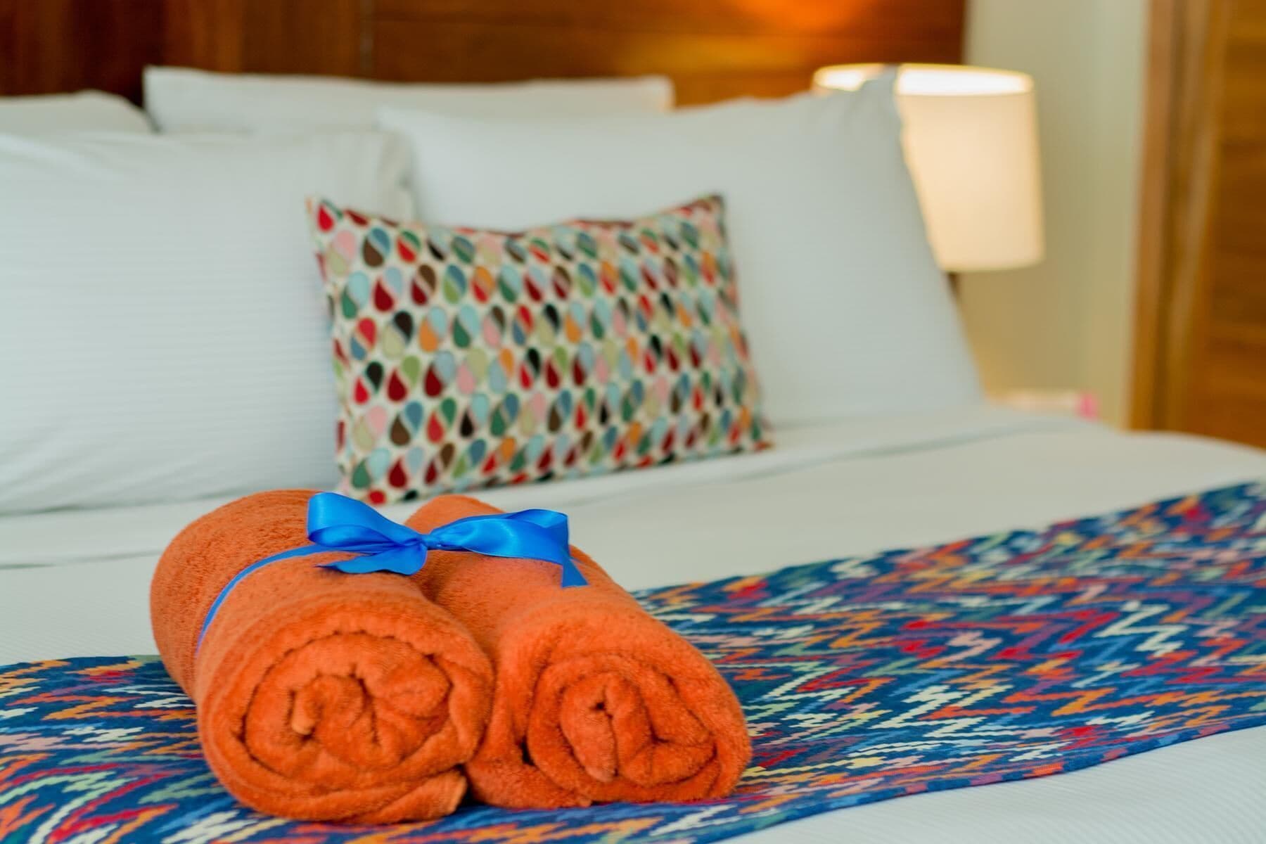 A neatly made bed with a patterned pillow and rolled towels with a blue ribbon.