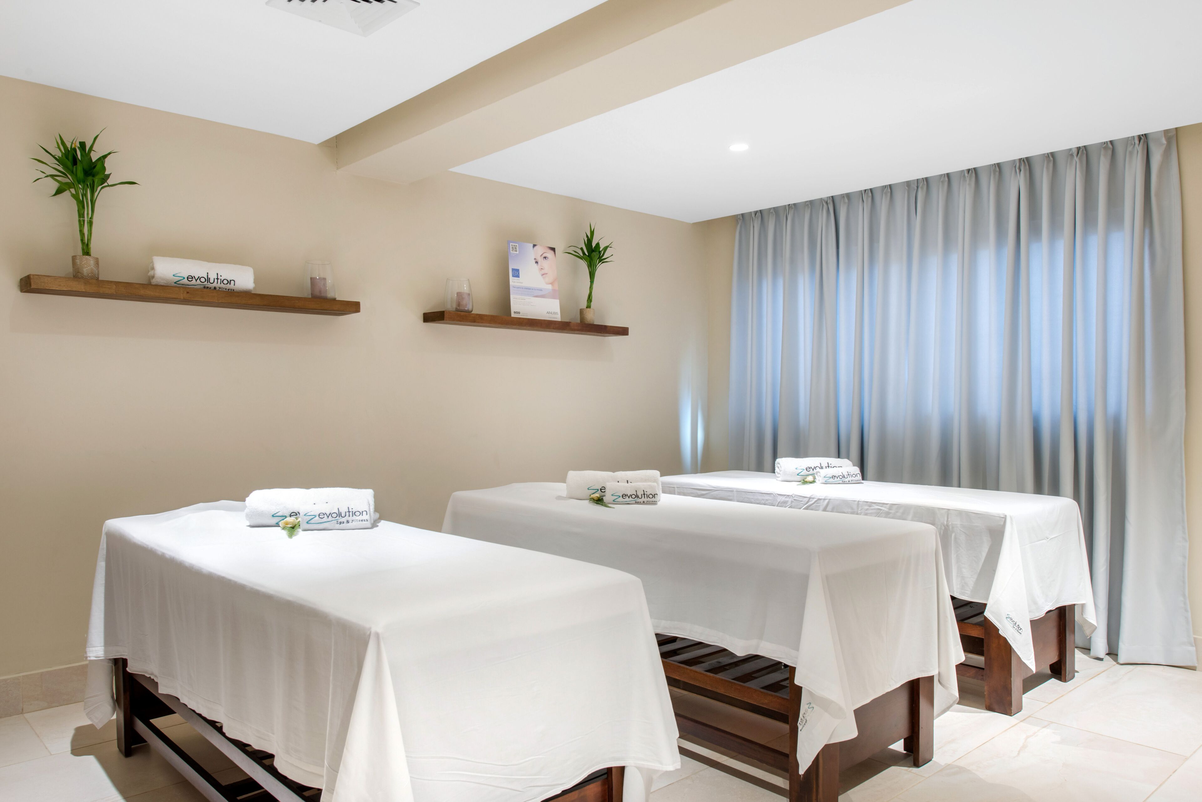 A spa room with two massage tables, each with a white tablecloth and a towel with 'evolution' branding. There are wooden shelves with plants and a framed picture on the wall.