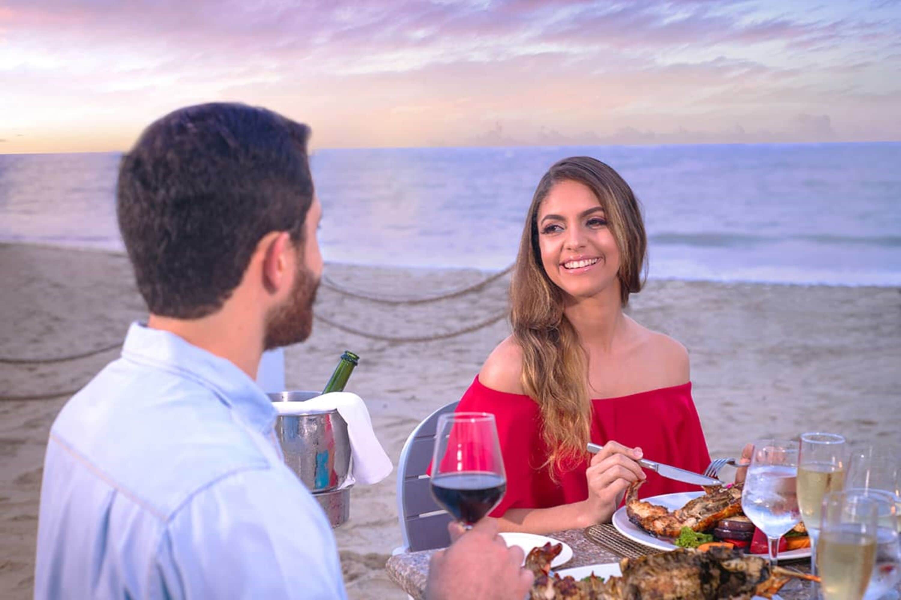 A couple dining at a table on a beach with the ocean in the background.