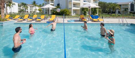 Una piscina con gente jugando, rodeada de sillones de descanso y sombrillas, con un edificio al fondo.
