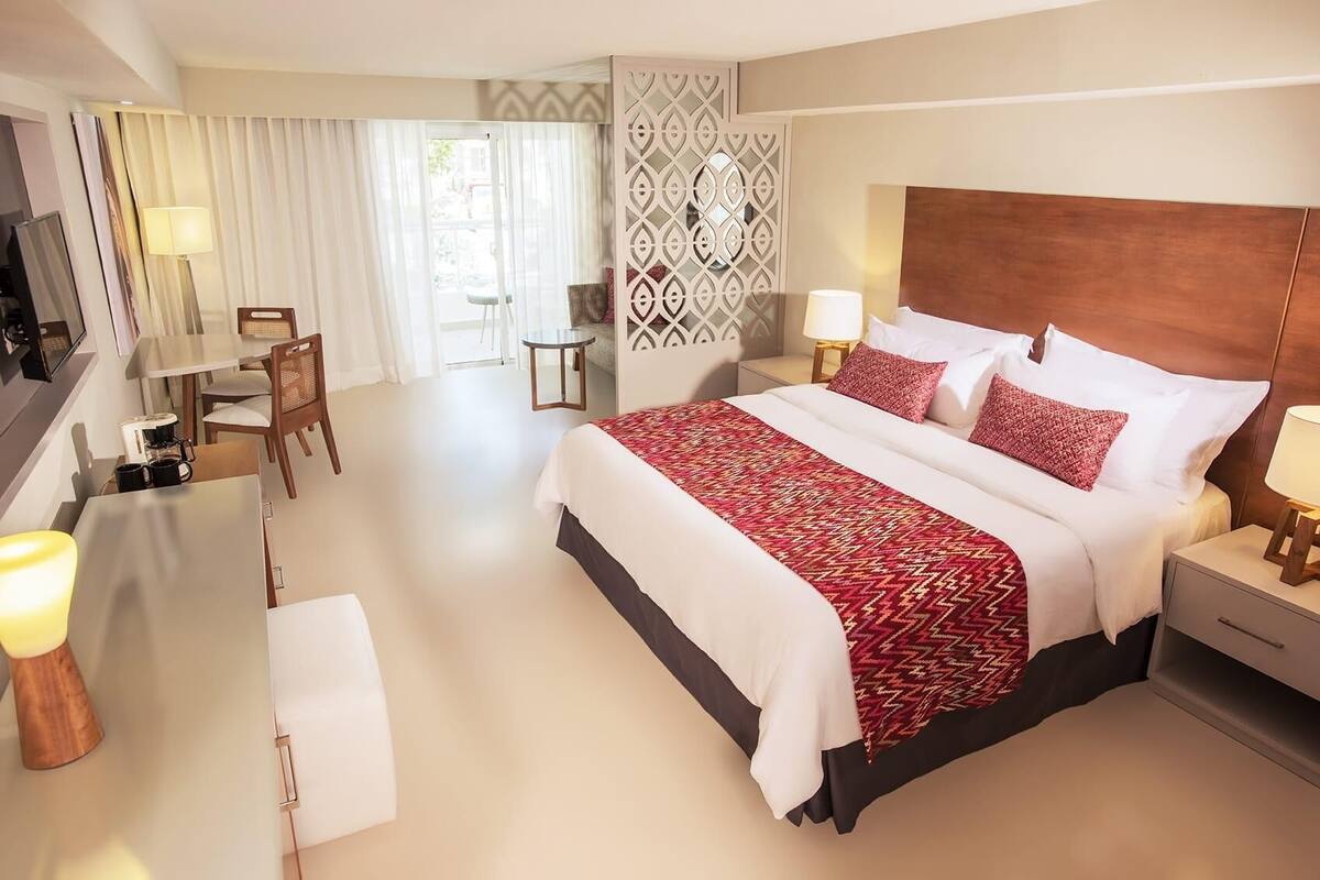 A hotel room with a large bed, a desk, a chair, and a dining area.
