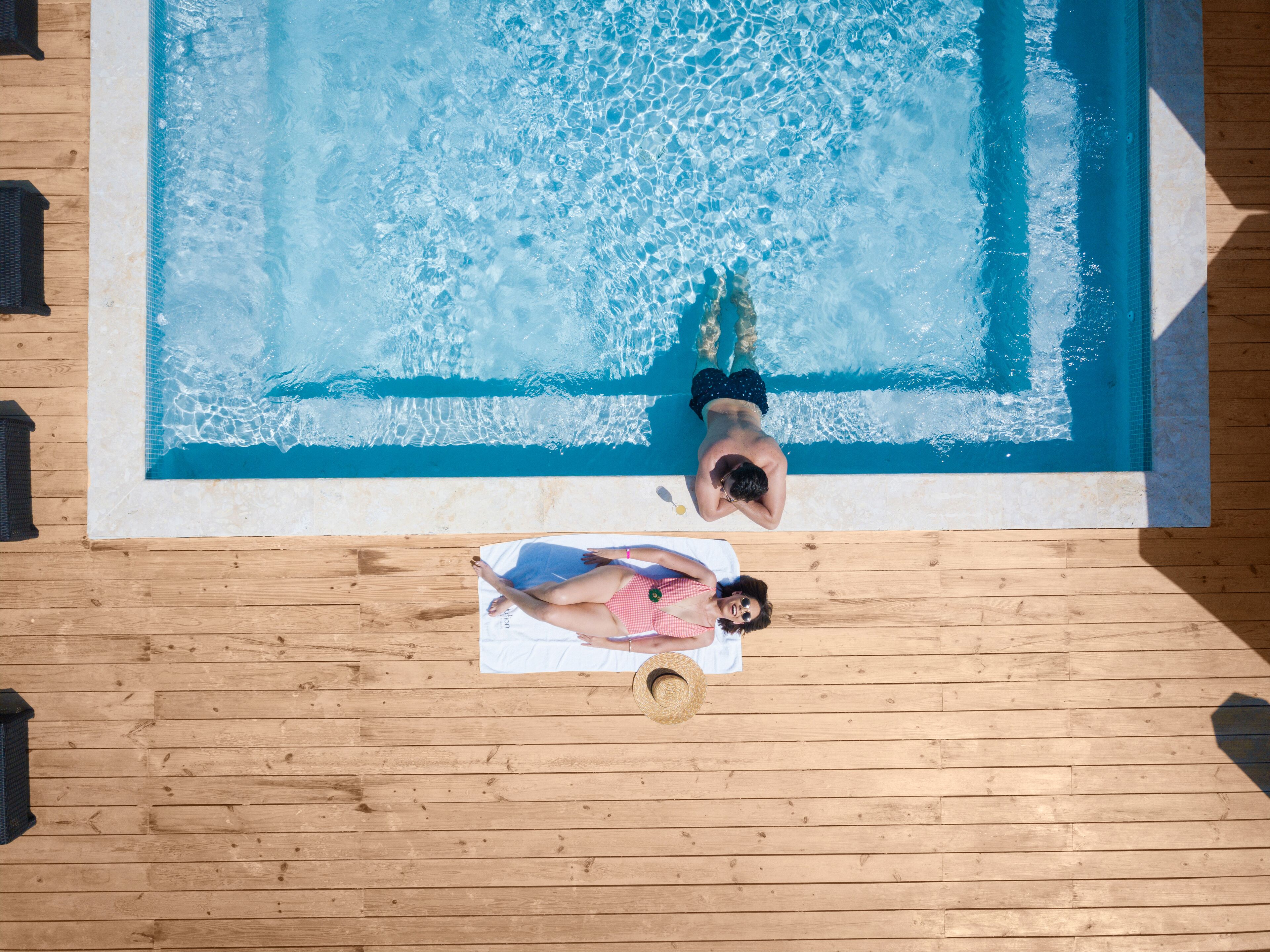 A swimming pool with two people sunbathing on a wooden deck.