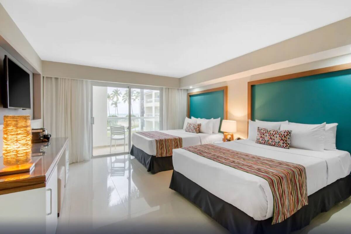 A hotel room with two beds, a TV, a large window, and a view of palm trees.
