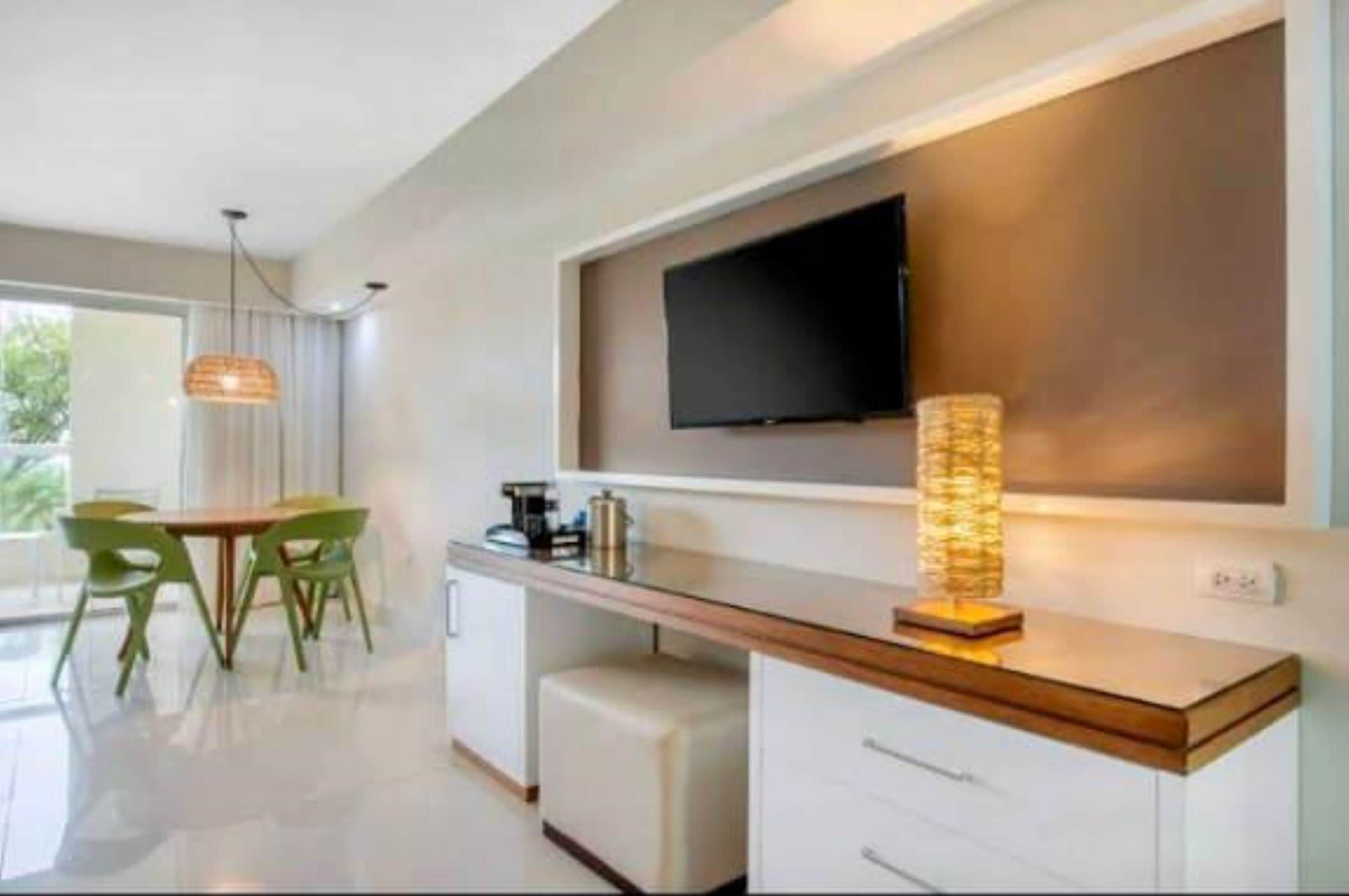 A modern hotel room with a flat-screen TV mounted on the wall, a wooden console table, a seating area with a table and chairs, and a large window providing natural light.