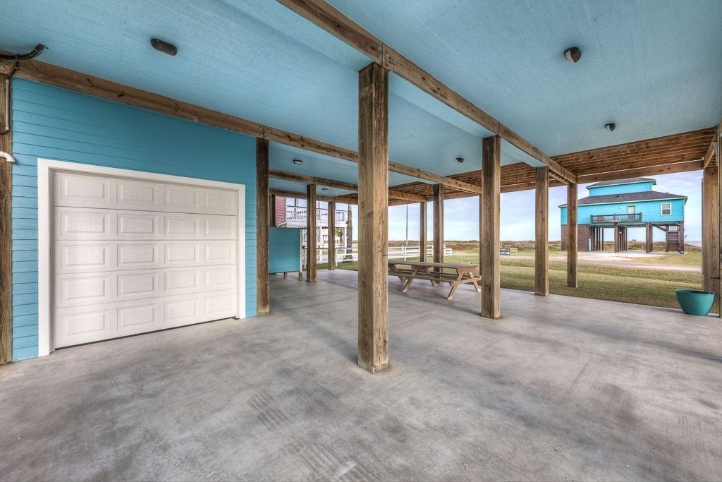 Un espace extérieur abrité, doté d’un sol en béton, de poutres en bois et d’une porte de garage blanche.