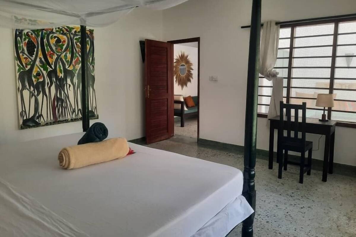 A hotel room with a bed, a desk, a chair, a window with curtains, a door, and a wall with a colorful painting.