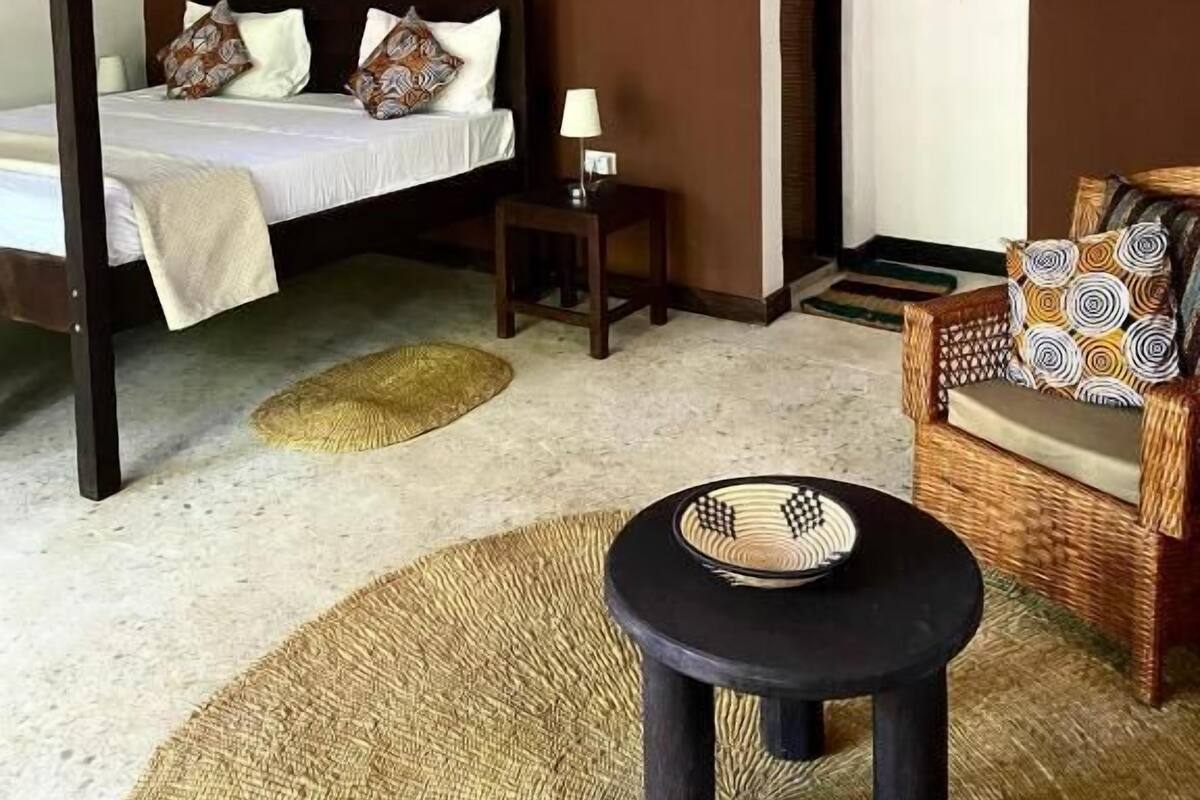 A bedroom with a four-poster bed, a small table with a lamp, a wicker chair, and a ceiling fan.