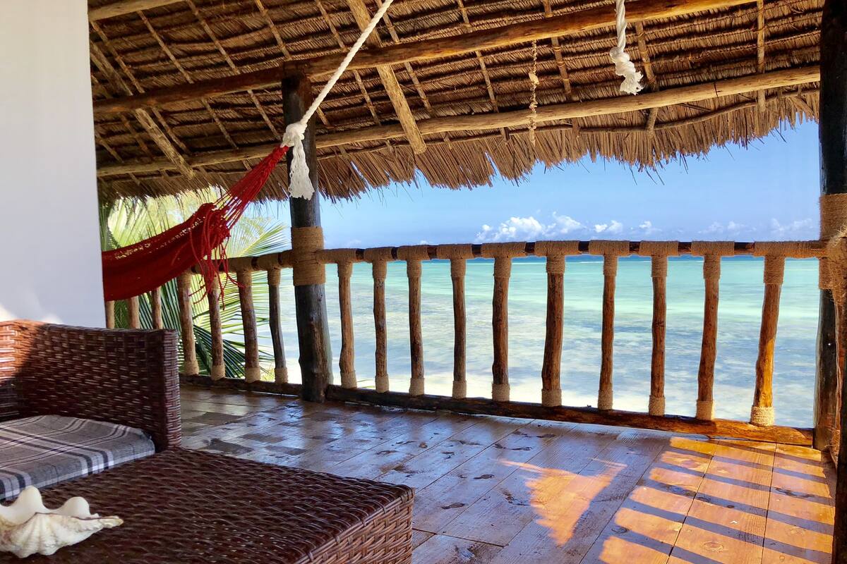 A wooden deck with a woven bench and a hammock, overlooking a beach with clear turquoise water.