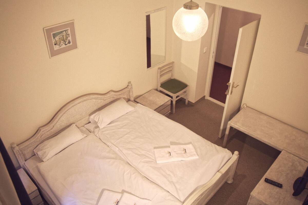 A neatly made bed with white linens, a bedside table with a remote control, a chair with a green cushion, and a mirror on the wall.