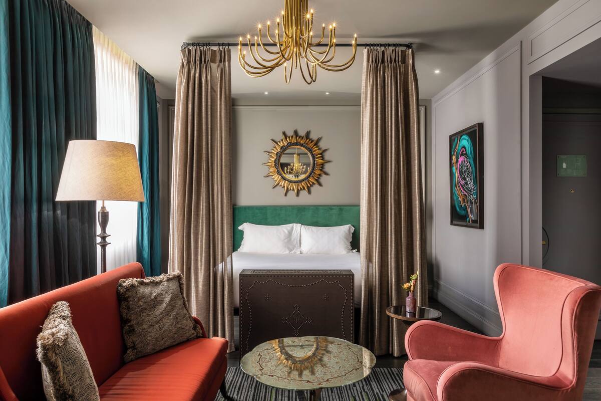 A hotel room with a bed, a red sofa, a pink armchair, a round coffee table, a chandelier, and a wall-mounted mirror.