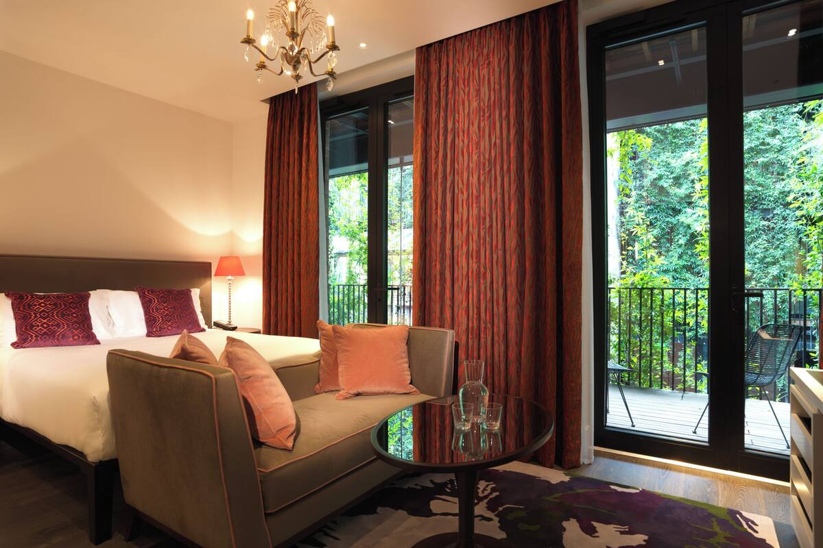 A hotel room with a bed, a sofa, a glass table, and a view of greenery outside.