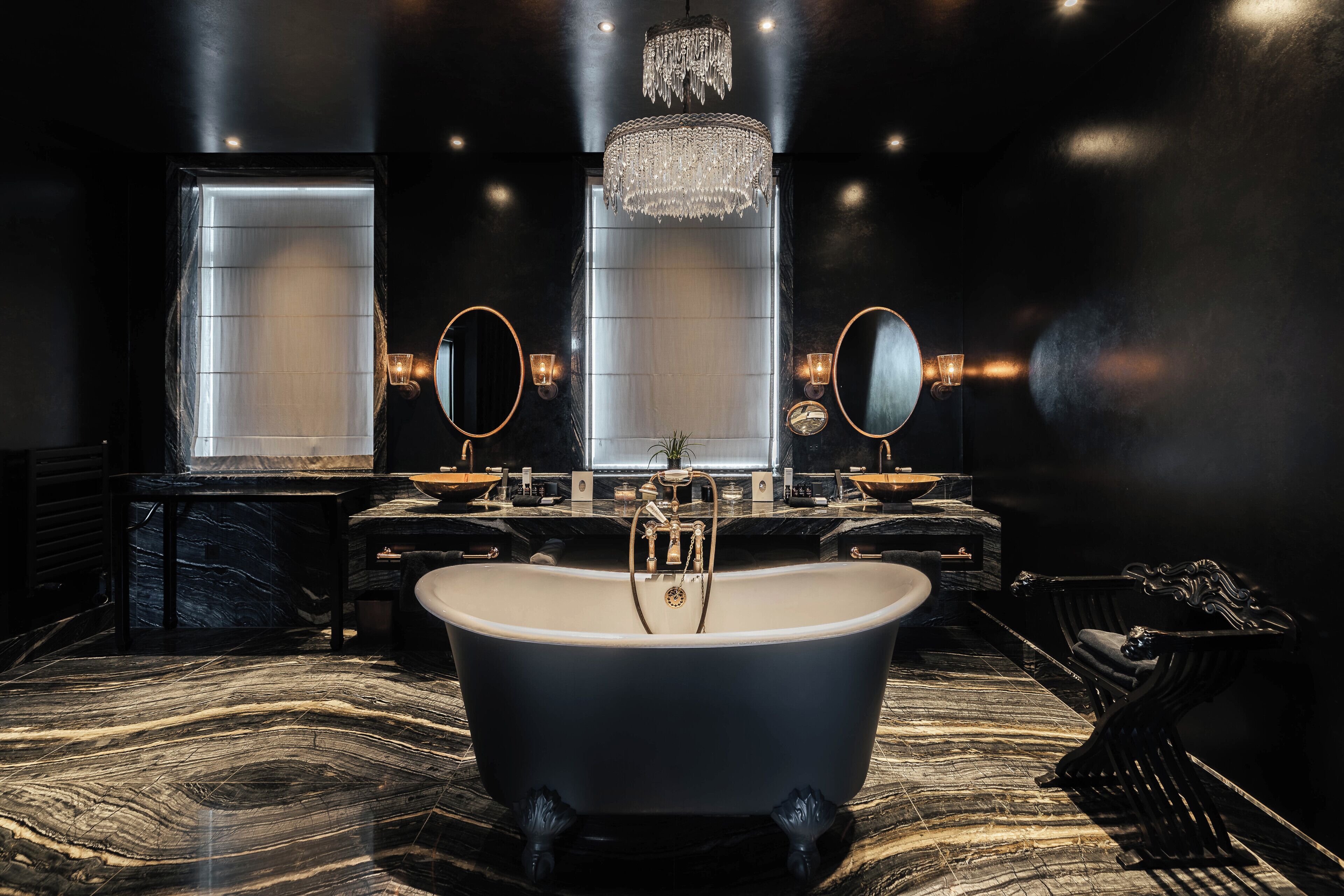 A luxurious bathroom with a freestanding bathtub, two round mirrors, a chandelier, and marble flooring.