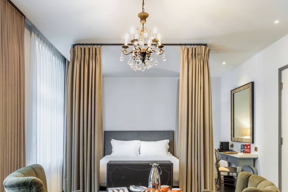 A hotel room with a bed, two armchairs, a desk, a chandelier, and a patterned rug.