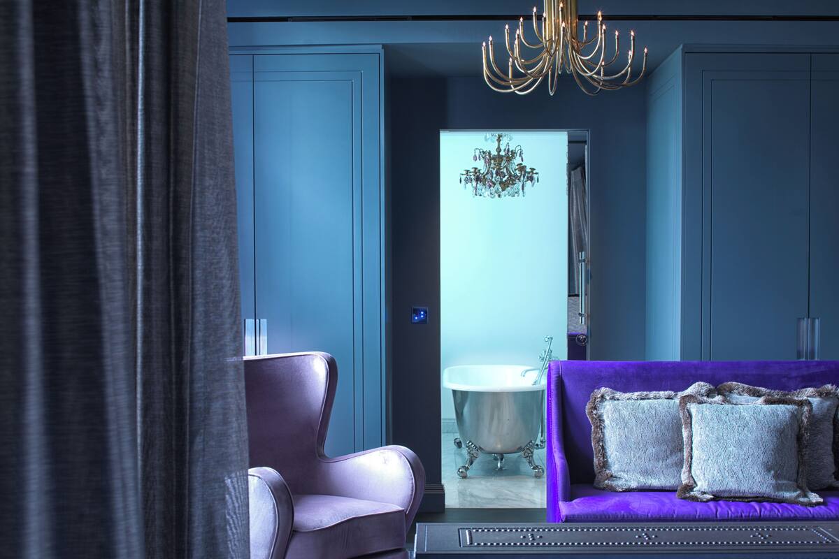 A modern bedroom with a purple sofa, a chair, a chandelier, and a bathtub.