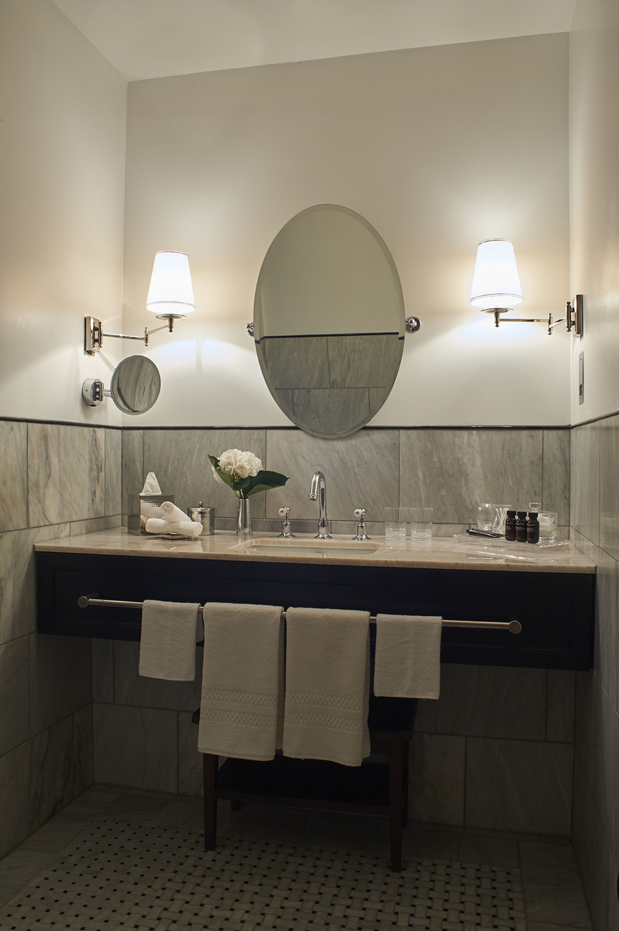 A bathroom with a marble vanity, two wall-mounted lamps, a round mirror, and a towel rack with towels.