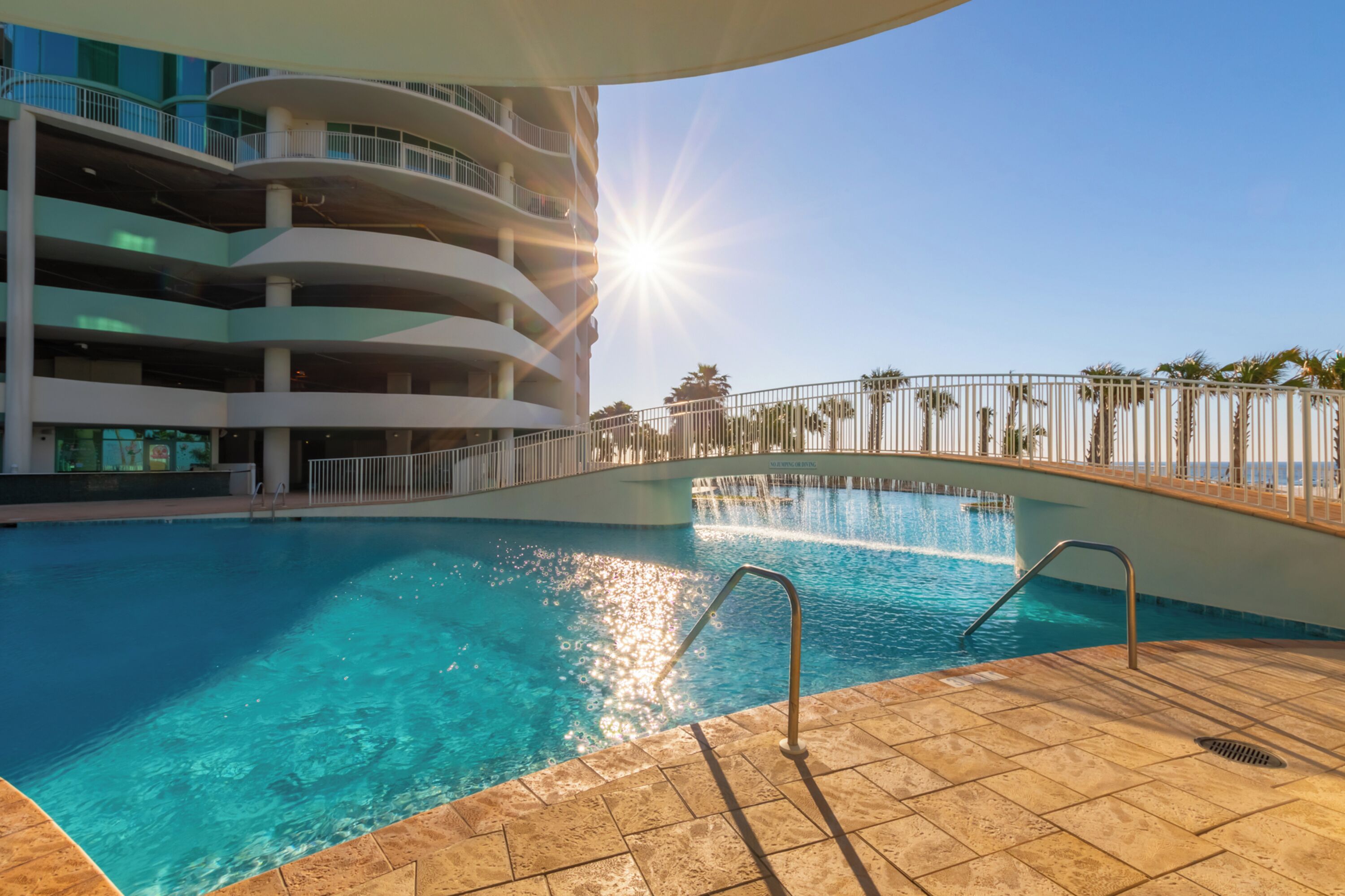 A swimming pool with a sun reflecting off the water, surrounded by a multi-level building with balconies.