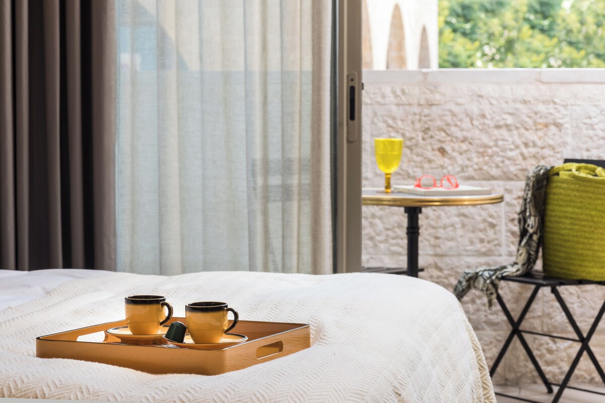 A hotel room with a bed, a tray with two cups, a glass of water, and a view of the outdoors.