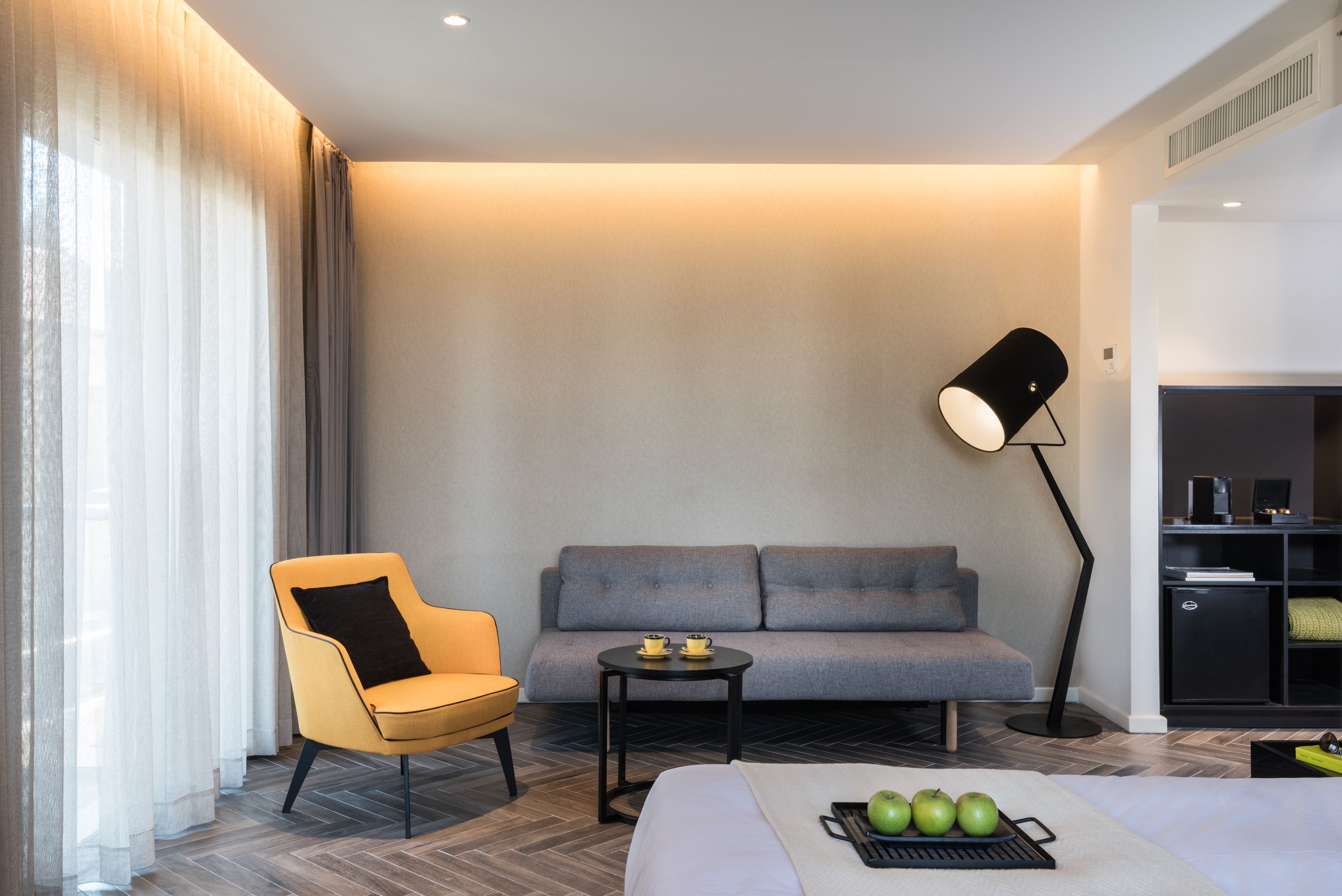 Ein modernes Hotelzimmer mit einer Couch, einem Sessel, einem kleinen Tisch mit Apfelschale, einer Lampe und einem Bett.