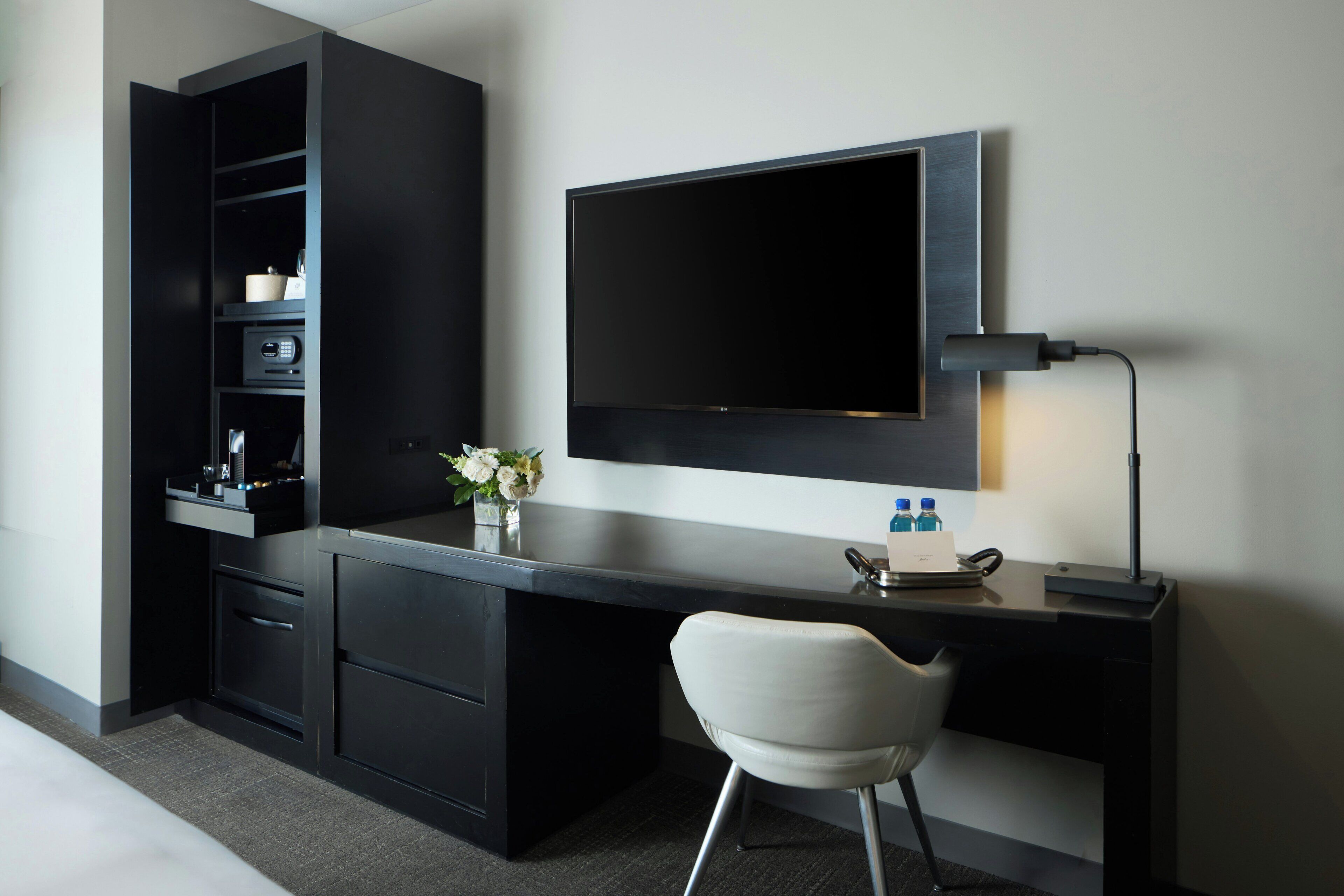 Habitación de hotel moderna con un escritorio oscuro, un televisor de pantalla plana montado encima y una silla blanca, junto con una mesita de noche con un florero.