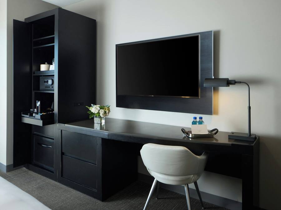 Habitación de hotel moderna con un escritorio oscuro, un televisor de pantalla plana montado encima y una silla blanca, junto con una mesita de noche con un florero.
