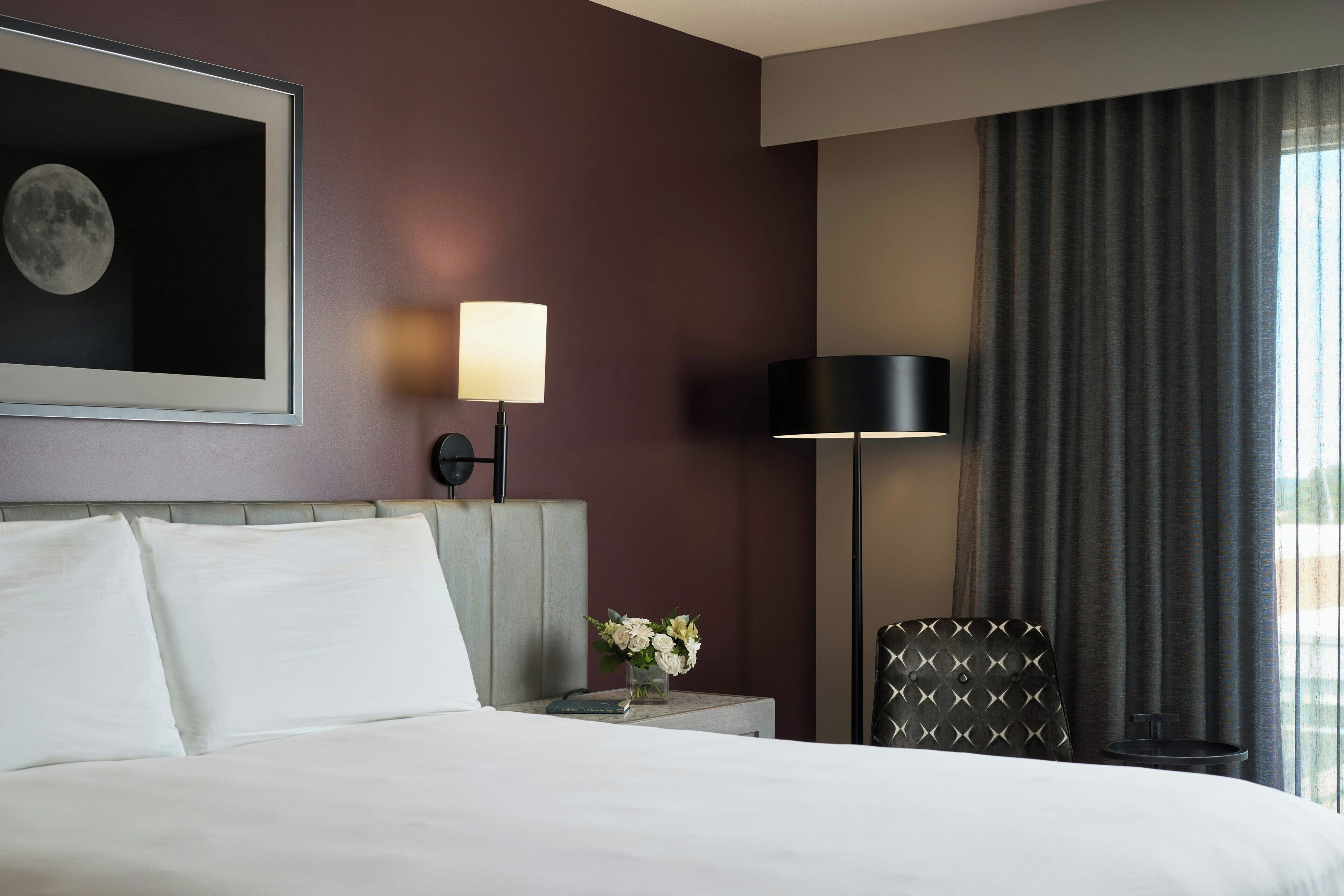 A hotel room with a bed, a nightstand with a lamp, a chair, and a window with curtains.