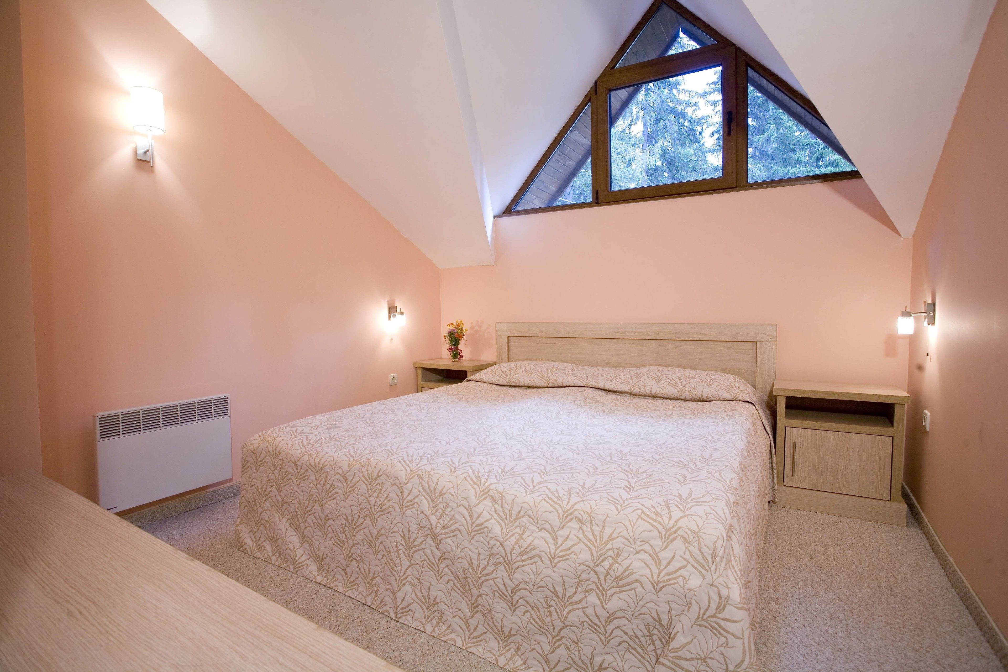 A bedroom with a sloped ceiling, a bed with a patterned bedspread, bedside lamps, a small nightstand, and a radiator.