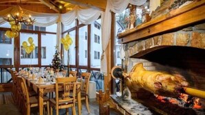 A dining area with a long wooden table set for a meal, a stone fireplace with a roasting pig, and a Christmas tree in the background. - Apart Hotel The Castle (Smolyan)