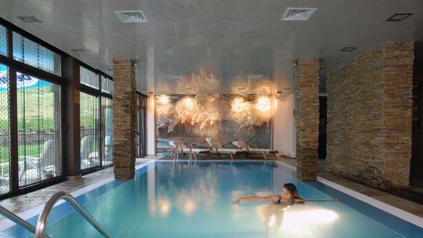 Una piscina interior con paredes de vidrio transparente, un mural de piedra y una escalera de piscina.