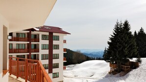 Un hotel de varios pisos con balcones, una ladera nevada y una montaña boscosa al fondo.
