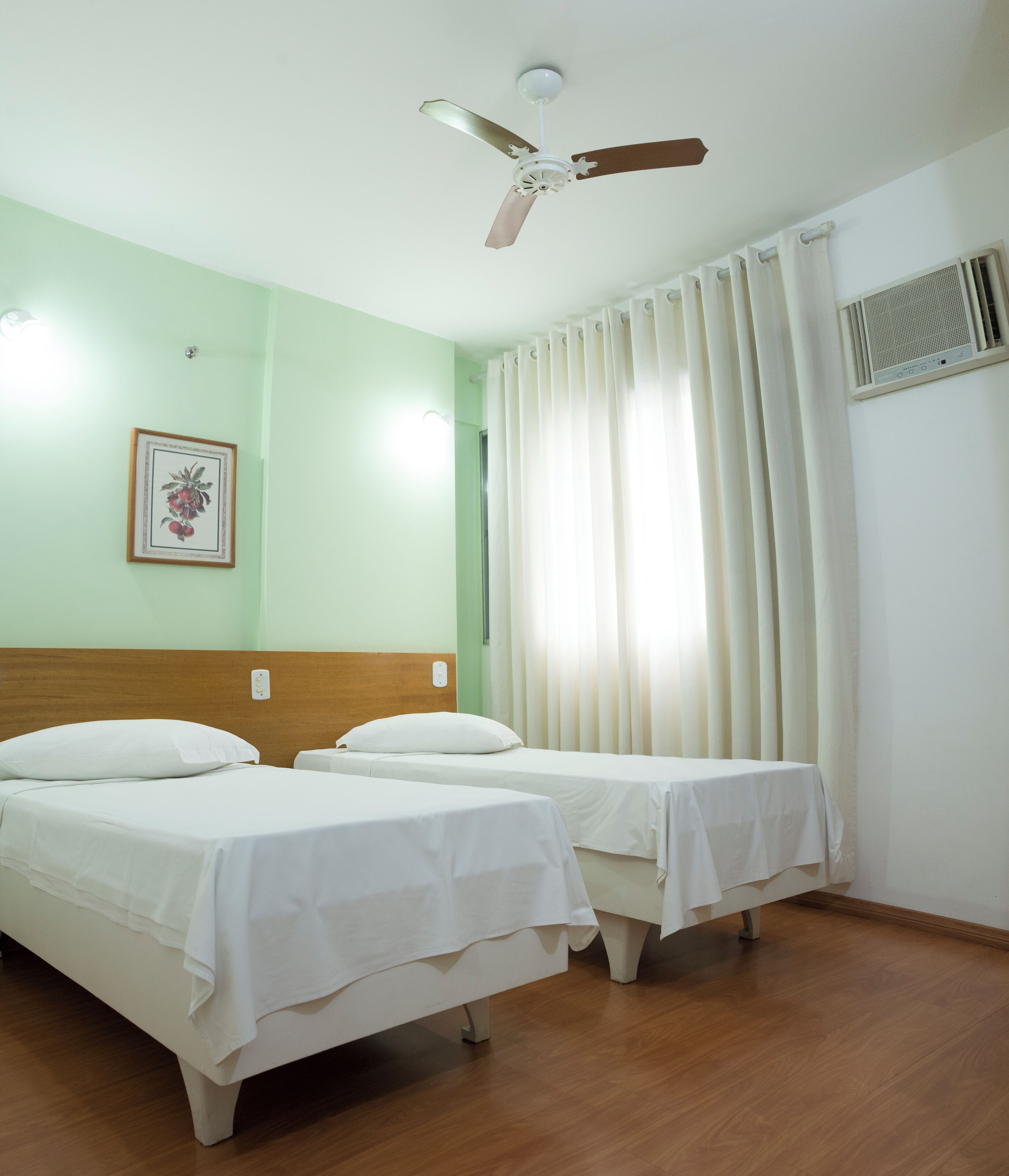 A hotel room with two single beds, a ceiling fan, a framed picture on the wall, and a window with curtains.