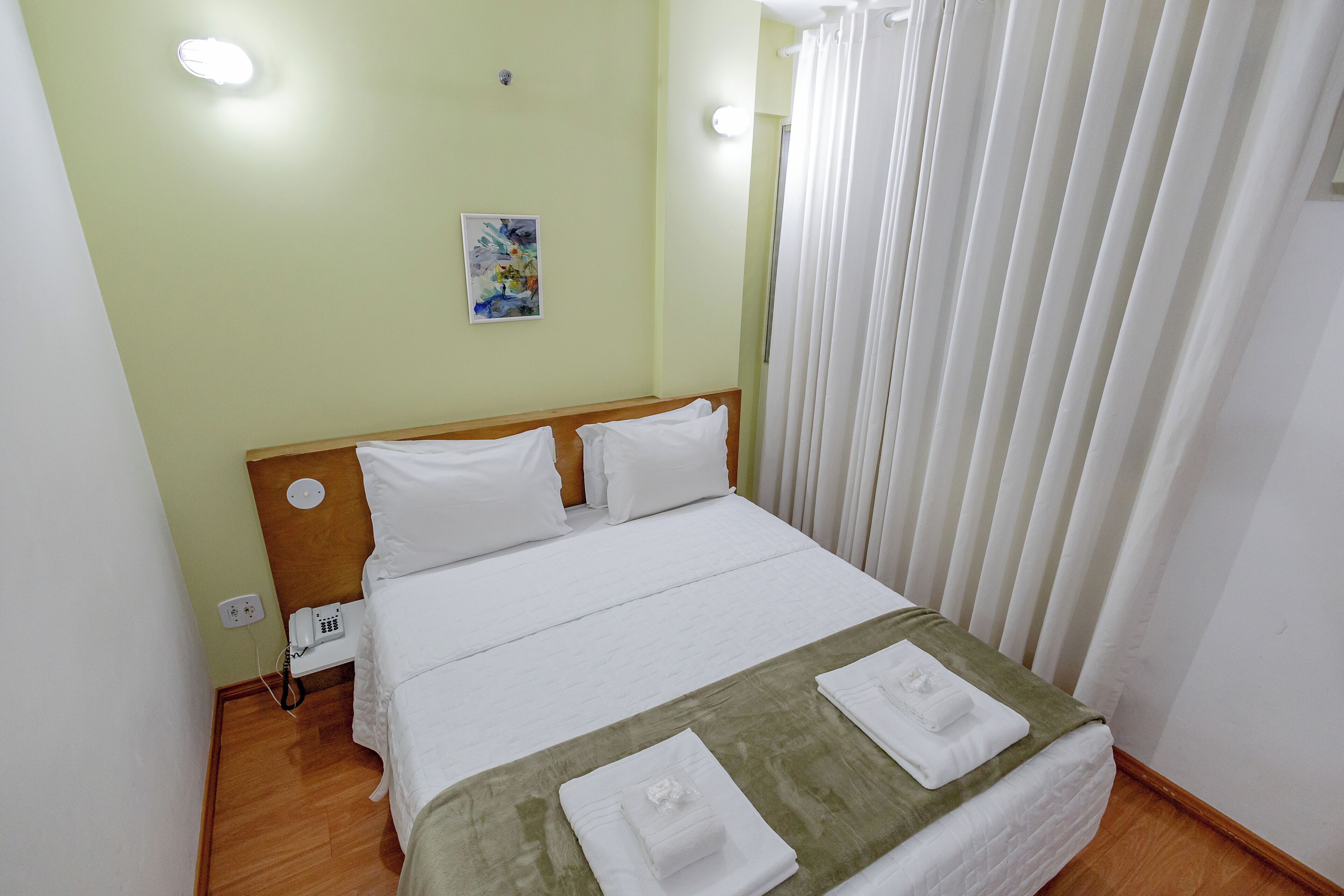 A neatly made bed with white linens, a green throw blanket, and two folded towels. A bedside table with a phone and a power outlet is visible. The room has light green walls and a window with white curtains.