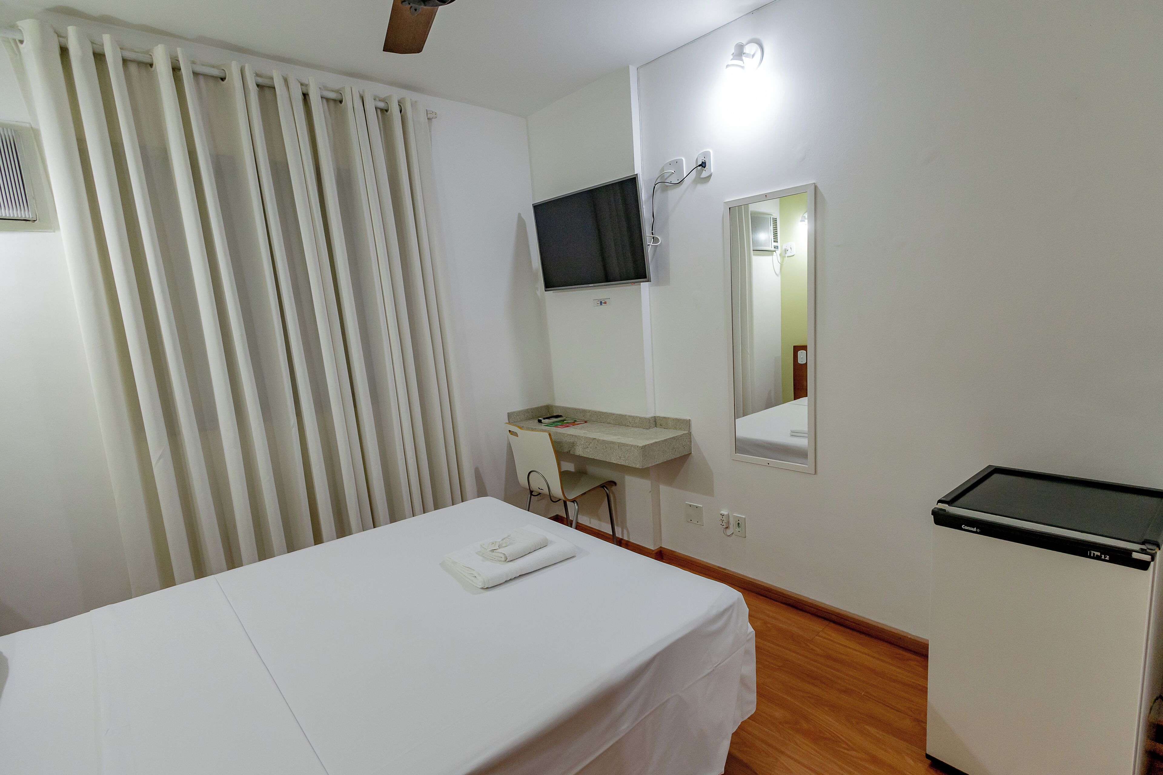 Una habitación de hotel con una cama, un escritorio, un televisor, un espejo y un refrigerador pequeño.